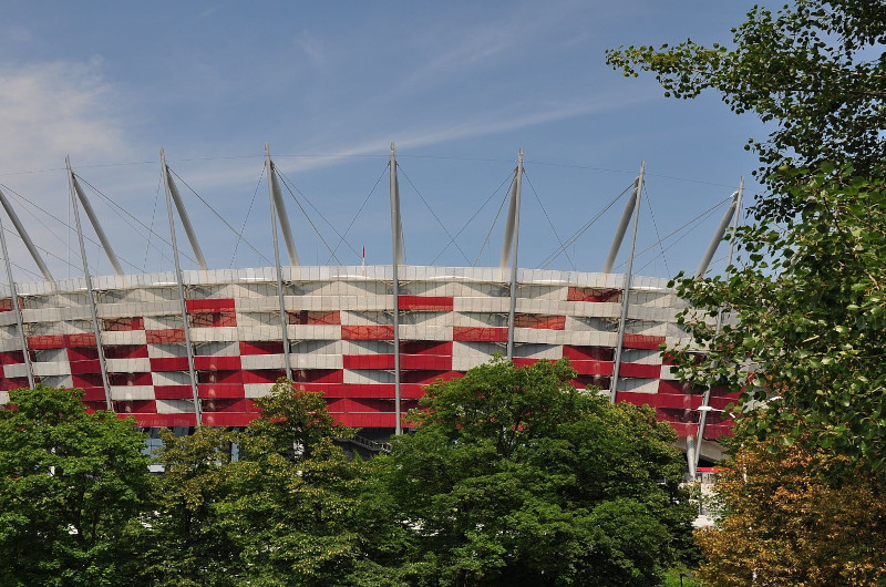 Obiekt sportowy: Stadion Narodowy z charakterystyczną elewacją w biało-czerwone pasy. Na górze wystają białe maszty z linami. Obiekt widoczny zza drzew.
