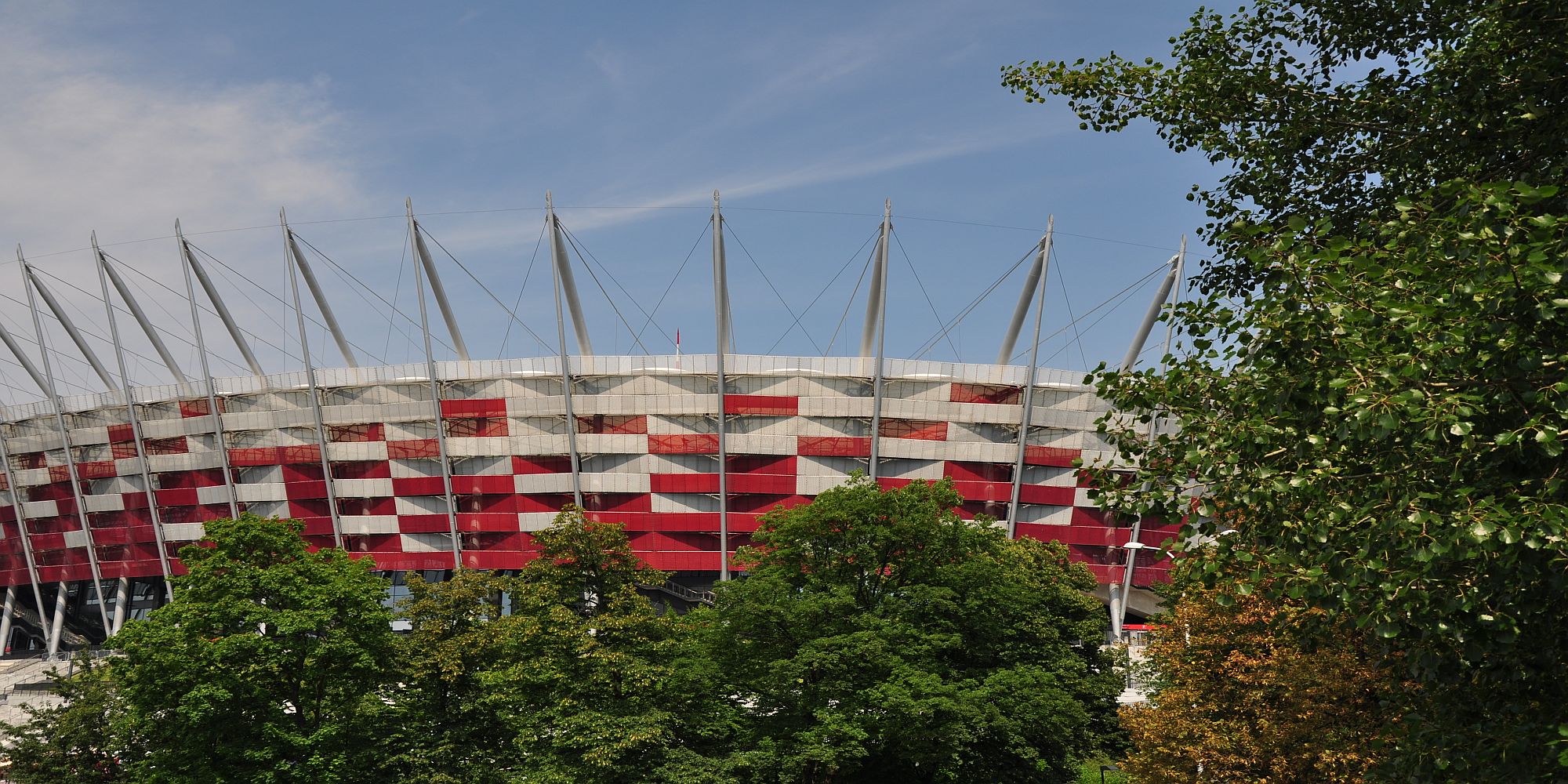 Obiekt sportowy: Stadion Narodowy z charakterystyczną elewacją w biało-czerwone pasy, widoczny zza drzew. Na górze wystają białe maszty z linami.