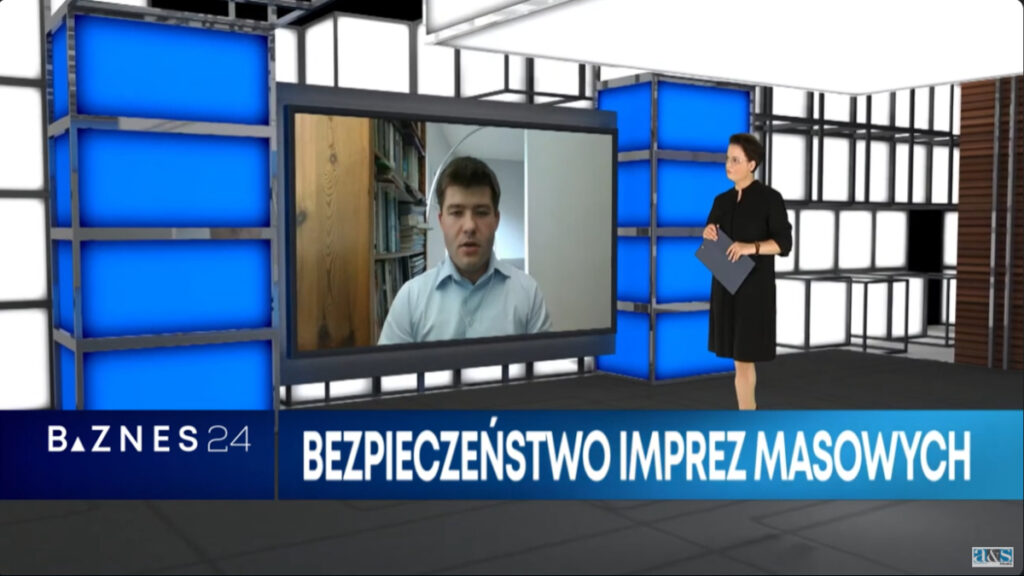 Studio telewizyjne. Kobieta w ciemnej sukience stoi na tle nowoczesnej scenografii z niebieskimi i białymi panelami. W rękach trzyma notatnik. Patrzy na duży ekran, na którym widoczny jest mężczyzna w jasnej koszuli uczestniczący w rozmowie zdalnie. Na dole biały napis na pasku w odcieniach niebieskiego: Bezpieczeństwo imprez masowych. W lewym dolnym rogu logo Biznes24.