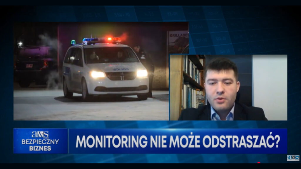 Po prawo okienko, a w nim mężczyzna ubrany w garnitur. Za nim półka z książkami. Po lewej radiowóz policyjny z włączonymi światłami na dachu. Na dole niebieski pasek z białym napisem: Monitoring nie może odstraszać? Obok napisu z lewej strony logo a&s i napis Bezpieczny Biznes. Tło czarno-granatowe.