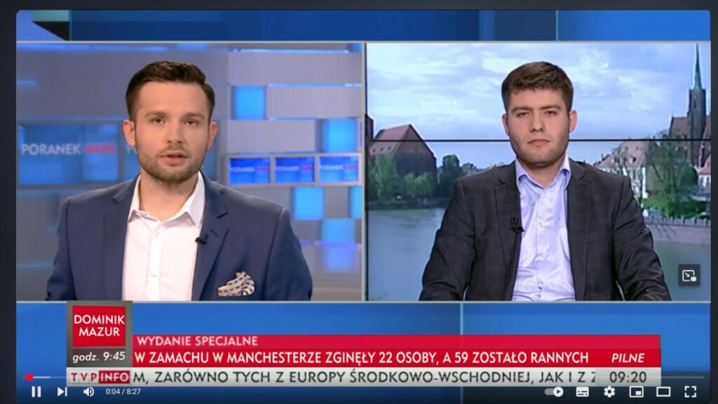 Dwóch mężczyzn w garniturach w studiu telewizyjnym podczas programu informacyjnego TVP Info Wydanie specjalne o zamachu bombowym w Manchesterze. Po lewej stronie dziennikarz, po prawej gość programu. Na dole pasek informacyjny z wiadomościami.